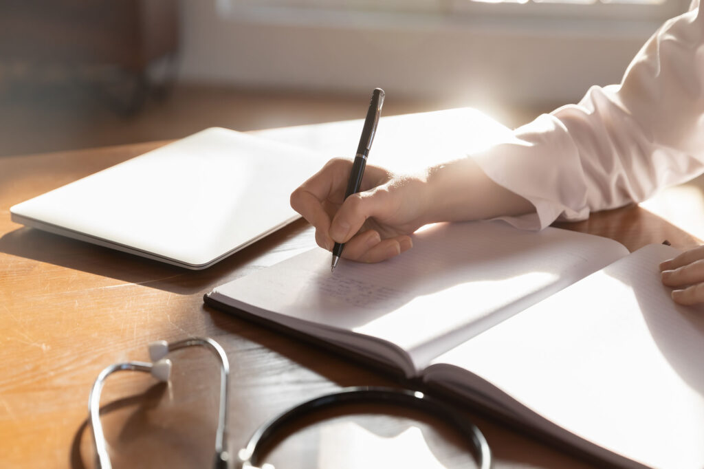 Close up woman doctor therapist writing in medical journal, sitting at table with stethoscope in office, working with documents, physician writing illness history, prescription in patient card